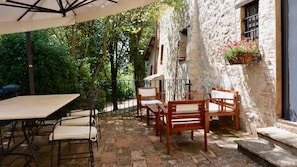 An outdoor patio with a table and chairs, a stone floor, and a rustic building with a window and flower box.