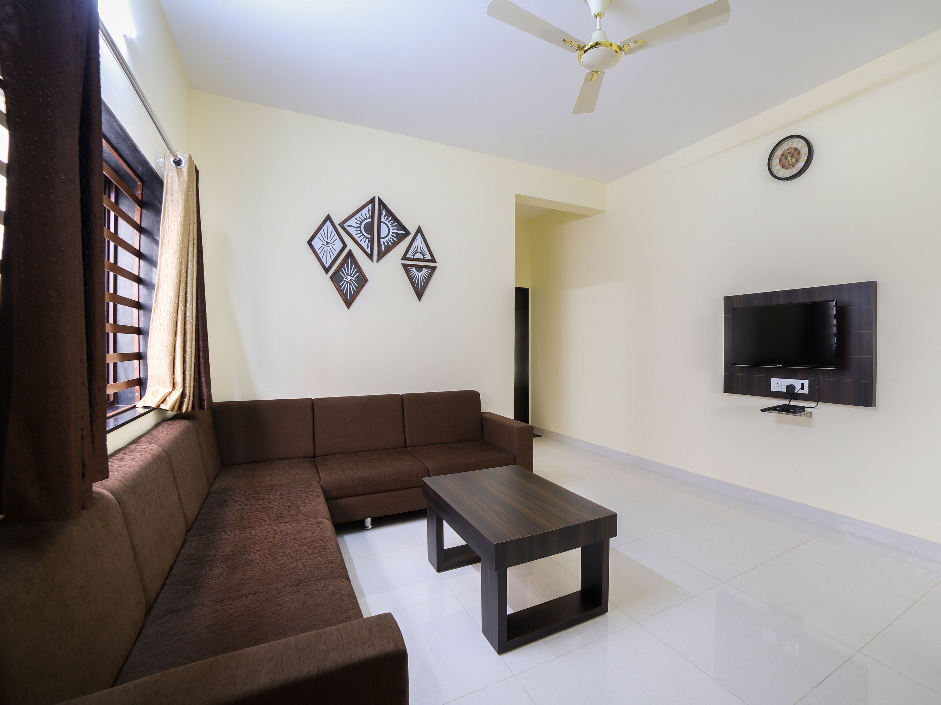 A living room with a brown sofa, a coffee table, a TV mounted on the wall, and a ceiling fan.