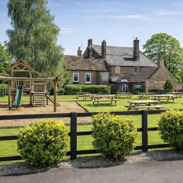A playground with a slide and climbing structure, picnic tables, and a fenced area with bushes. - The Bell Inn (Gloucester)