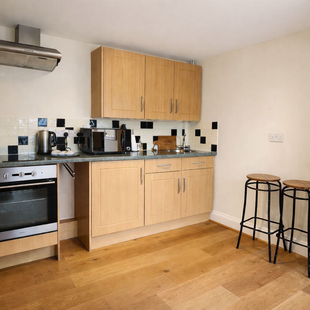 A modern kitchen with wooden cabinets, a stainless steel range hood, a microwave, a kettle, and two stools.