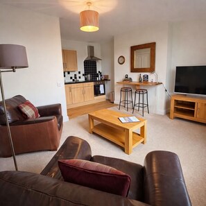 A living room with a sofa, a coffee table, a TV, and a kitchen in the background.