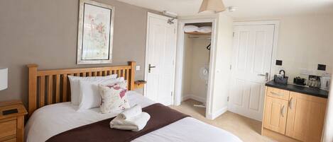 A bedroom with a large bed, wooden headboard, a bedside table, a nightstand, a wardrobe, and a small kitchenette area.