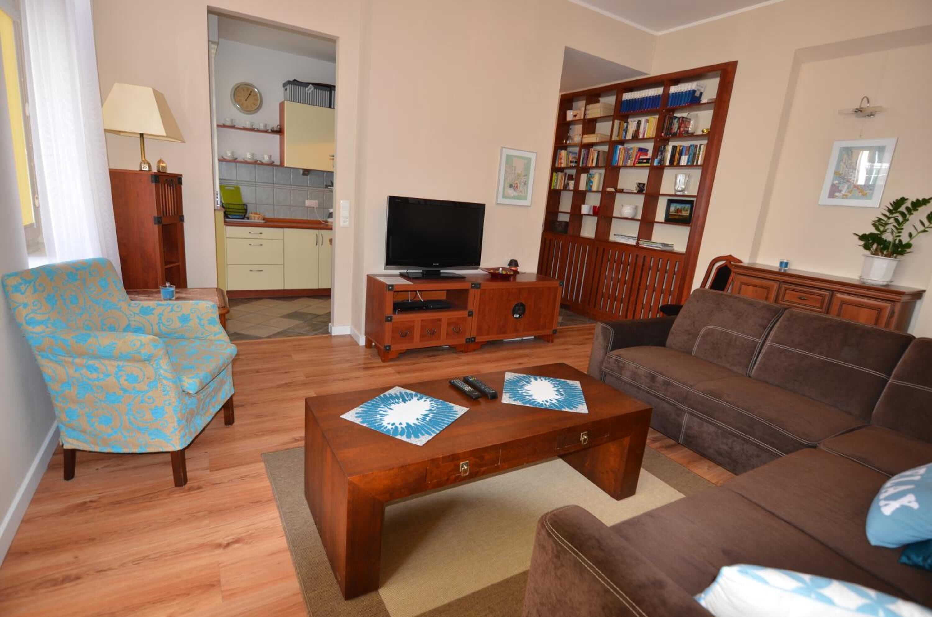 A living room with a sofa, armchair, coffee table, TV, bookshelf, and a kitchen in the background.