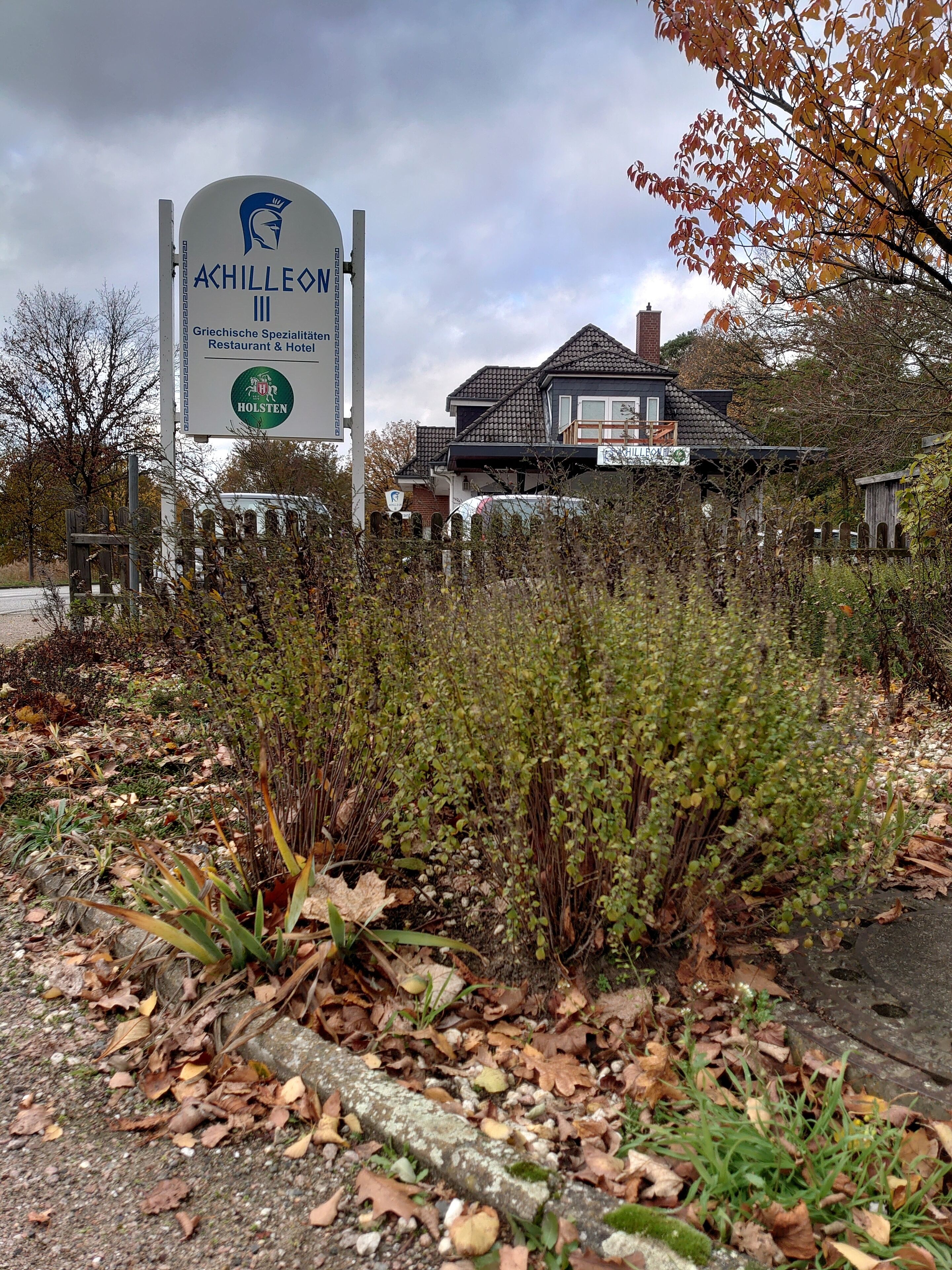 Une enseigne pour le restaurant et l’hôtel « Achilleo III », avec un logo, entourée de feuillage d’automne et d’une clôture.