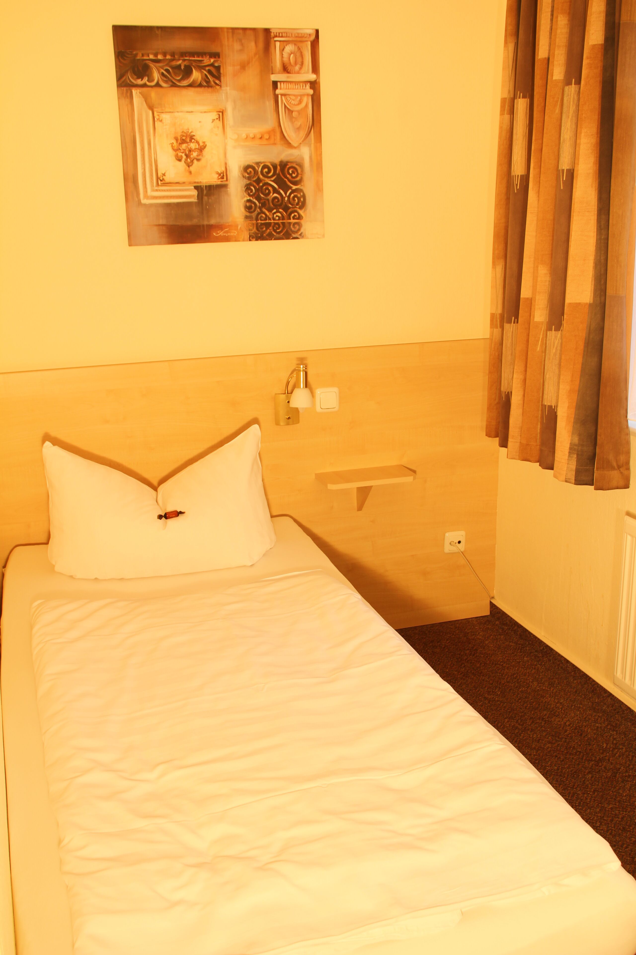 A hotel room with a bed, a nightstand, a wall-mounted lamp, and a painting above the bed.