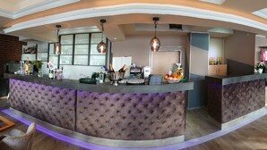 A bar area with a tufted bar counter, hanging pendant lights, and a variety of bottles and glasses. - Achilleon III (Geesthacht)