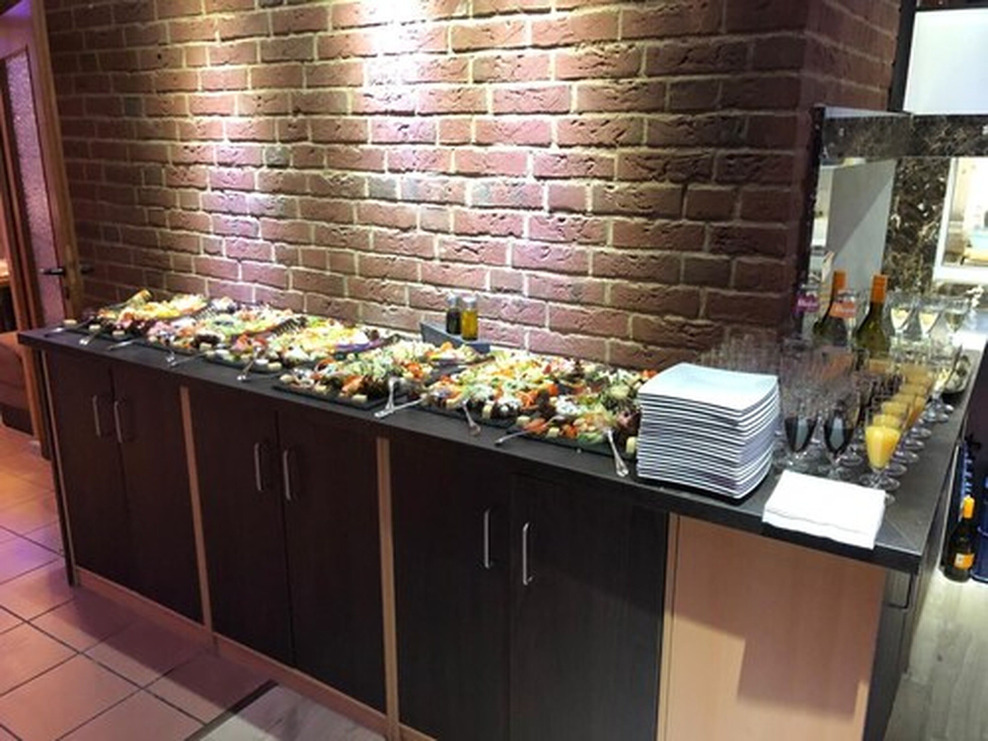 A buffet setup with various food items and a stack of plates on a counter against a brick wall.