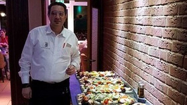 A buffet setup with a variety of dishes and drinks, a stack of plates, and a person standing behind the buffet. - Achilleon III (Geesthacht)