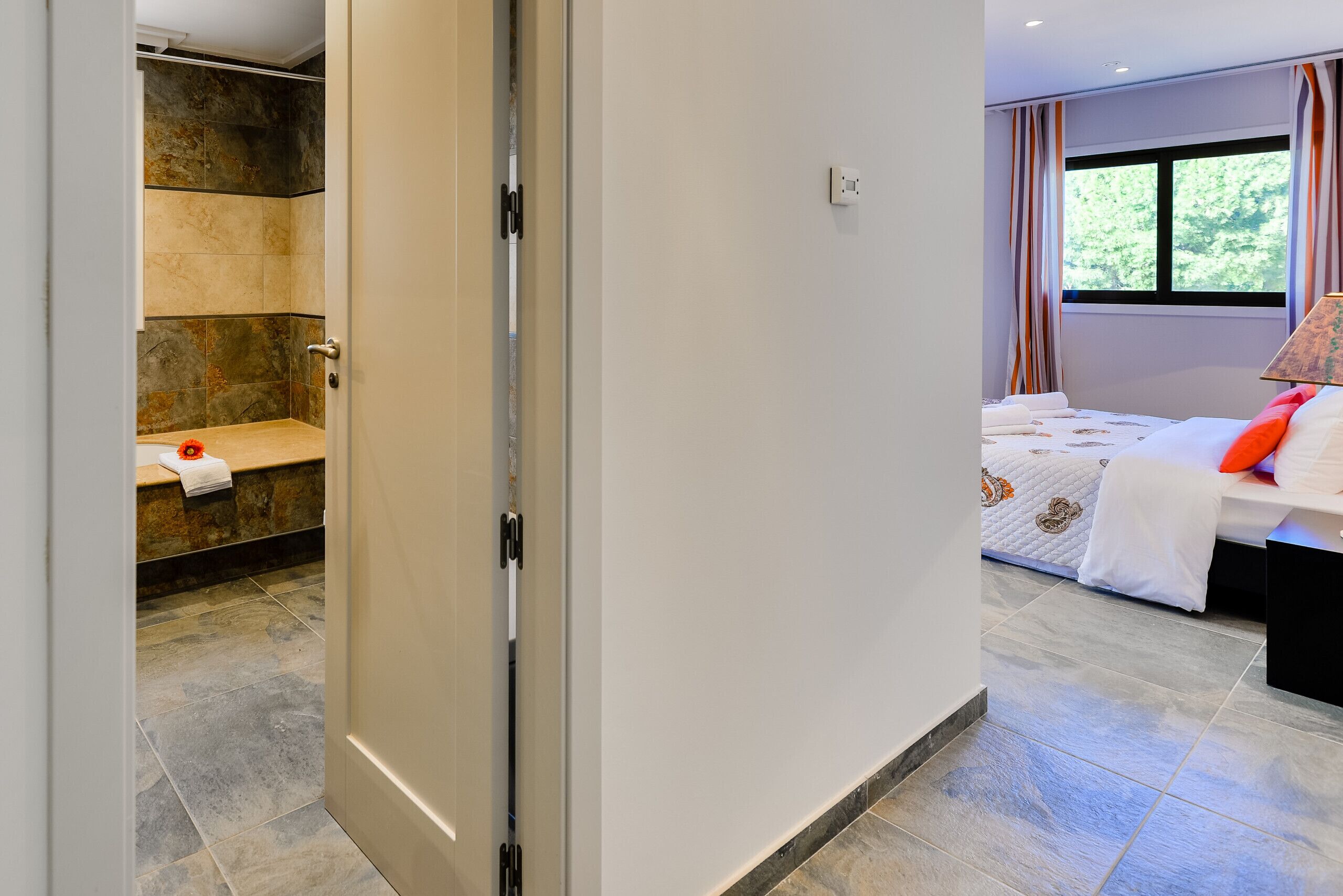 Une chambre d’hôtel moderne dotée d’une cabine de douche, d’un lit recouvert d’une courtepointe blanche et orange, et d’une fenêtre avec des rideaux.