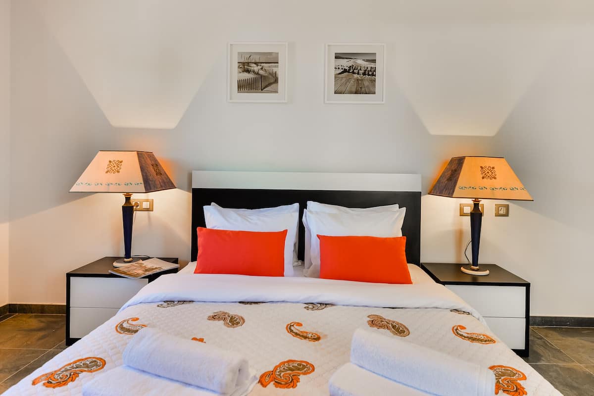 A neatly made bed with white and orange bedding, two nightstands with lamps, and two framed pictures on the wall.