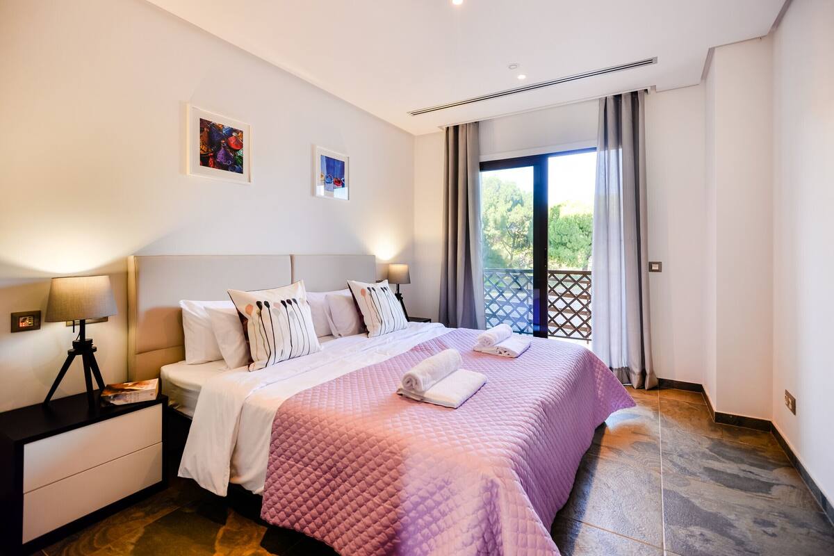 A bedroom with a large bed, a nightstand, a lamp, and a view of the outdoors through a window.
