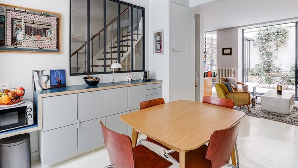A modern kitchen with a large window, a dining area with a wooden table and chairs, and a staircase leading to an upper floor. - Sumptuous house with patio (Paris)