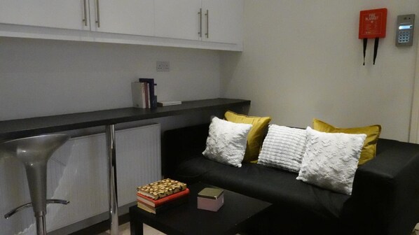 A modern living room with a black sofa, white and yellow cushions, a small coffee table, and a radiator. - Grand Airport Ensuites (West Drayton)
