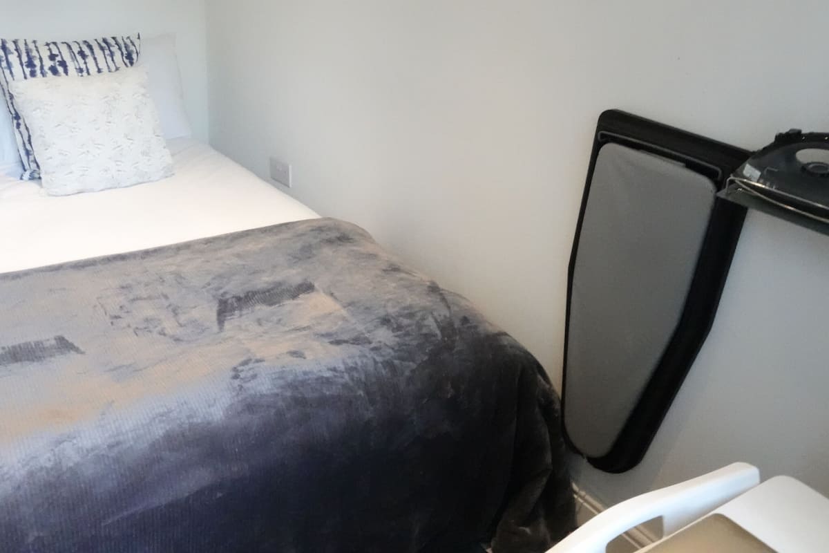 Ein Bett mit dunkler Decke, einem weißen Kissen und einem gemusterten Kissen. An der Wand befindet sich ein schwarzes Objekt, möglicherweise eine Heizung oder Klimaanlage.