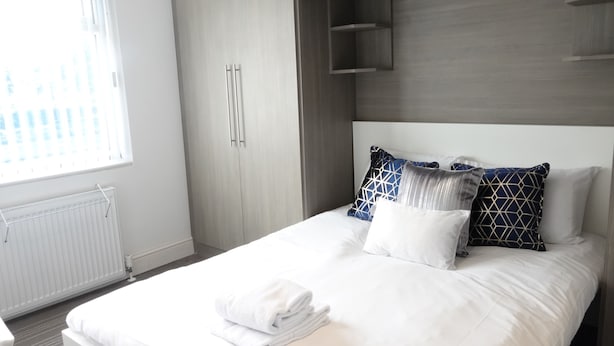 A neatly made bed with white linens and a folded towel, a wooden wardrobe, and a window with blinds. - Grand Airport Ensuites (West Drayton)