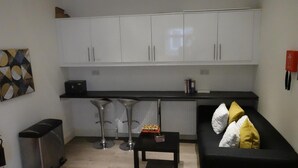 A compact living space with a kitchenette, a black sofa with cushions, a small table, and a bar stool. - Grand Airport Ensuites (West Drayton)