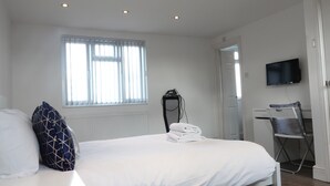 A hotel room with a bed, a window with blinds, a television mounted on the wall, and a chair. - Grand Airport Ensuites (West Drayton)