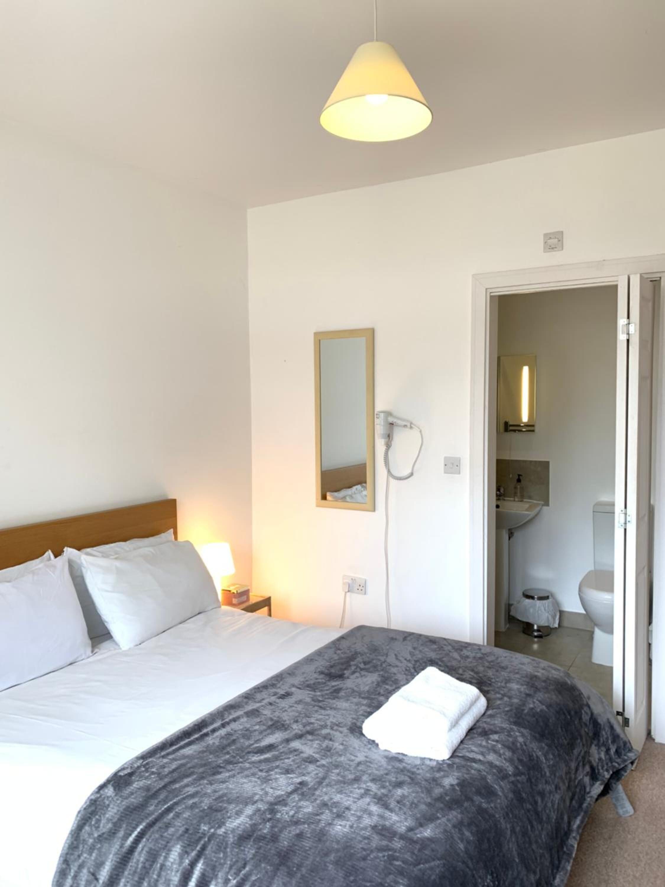 Camera d'albergo con un letto, una lampada da comodino, uno specchio e un bagno parzialmente visibile attraverso una porta aperta.