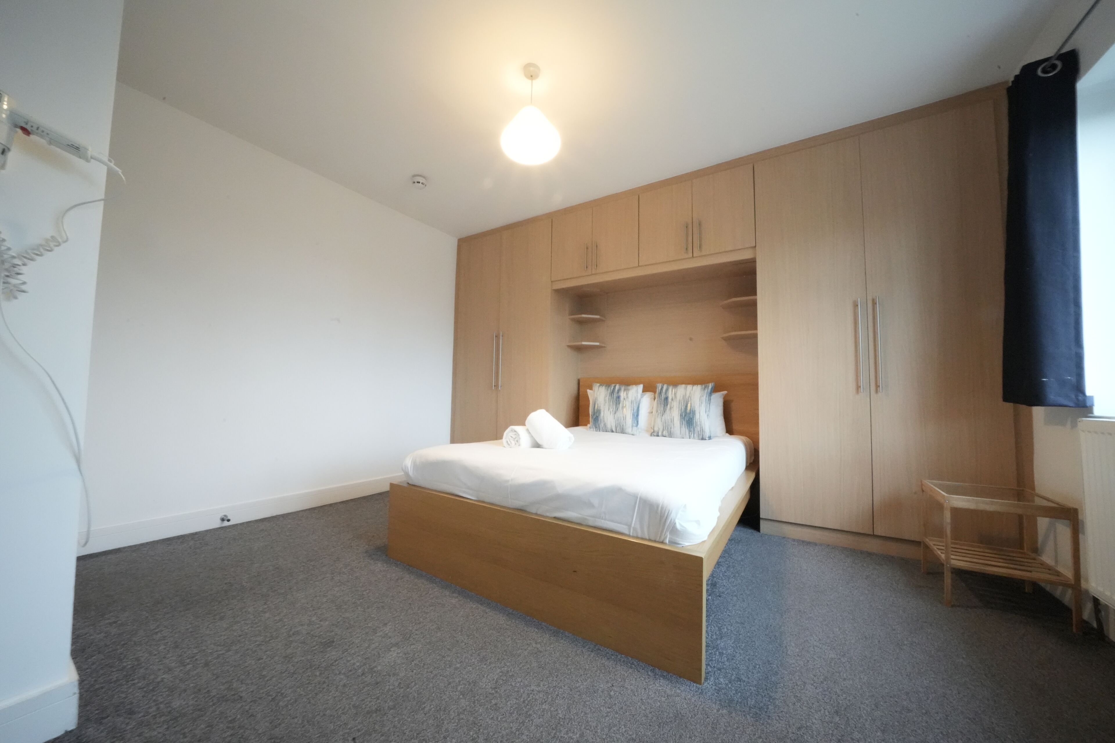 A bedroom with a wooden bed, grey carpet, a small wooden table, and built-in wooden cabinets.