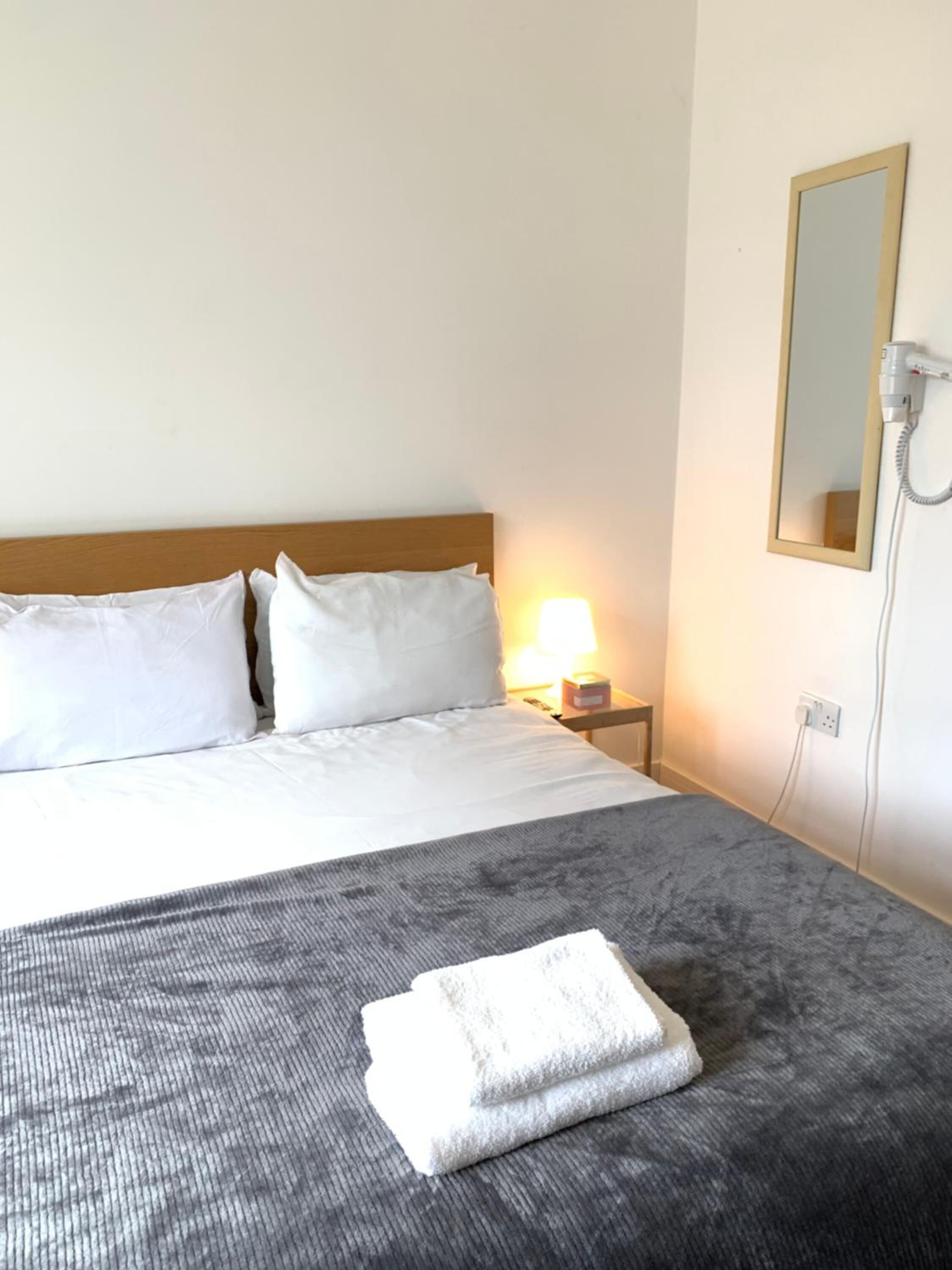A hotel room with a bed, a nightstand, a lamp, a mirror, and a folded towel on the bed.