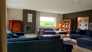 A modern living room with dark sofas, a fireplace, a bookshelf, and a large window. - A spacious and quiet villa in the heart of Flanders, near Bruges and the sea! (Maldegem)