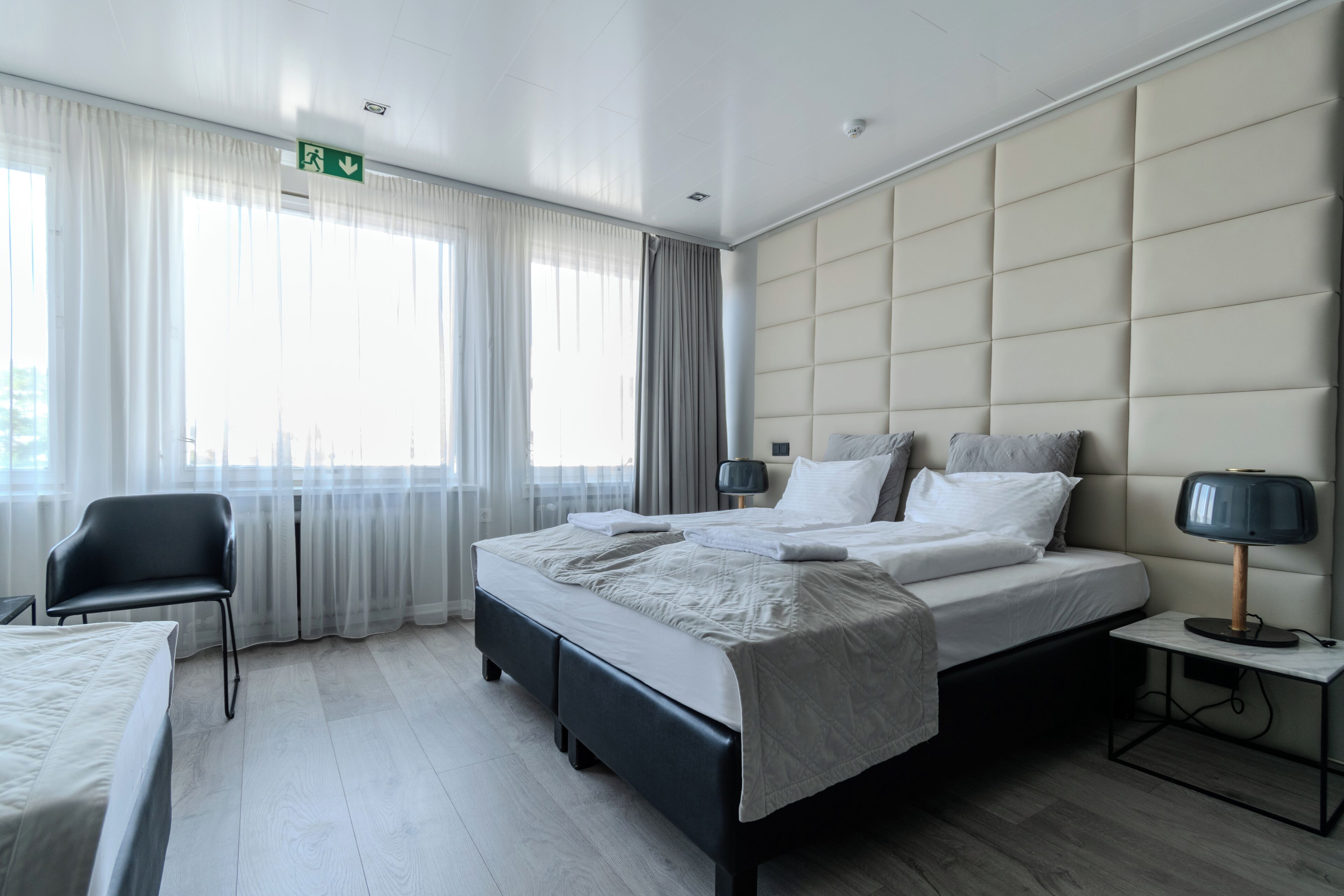 Une chambre d’hôtel moderne, dotée d’un grand lit, d’une chaise, d’une table de chevet avec une lampe et offrant une vue sur la ville grâce à de grandes fenêtres.