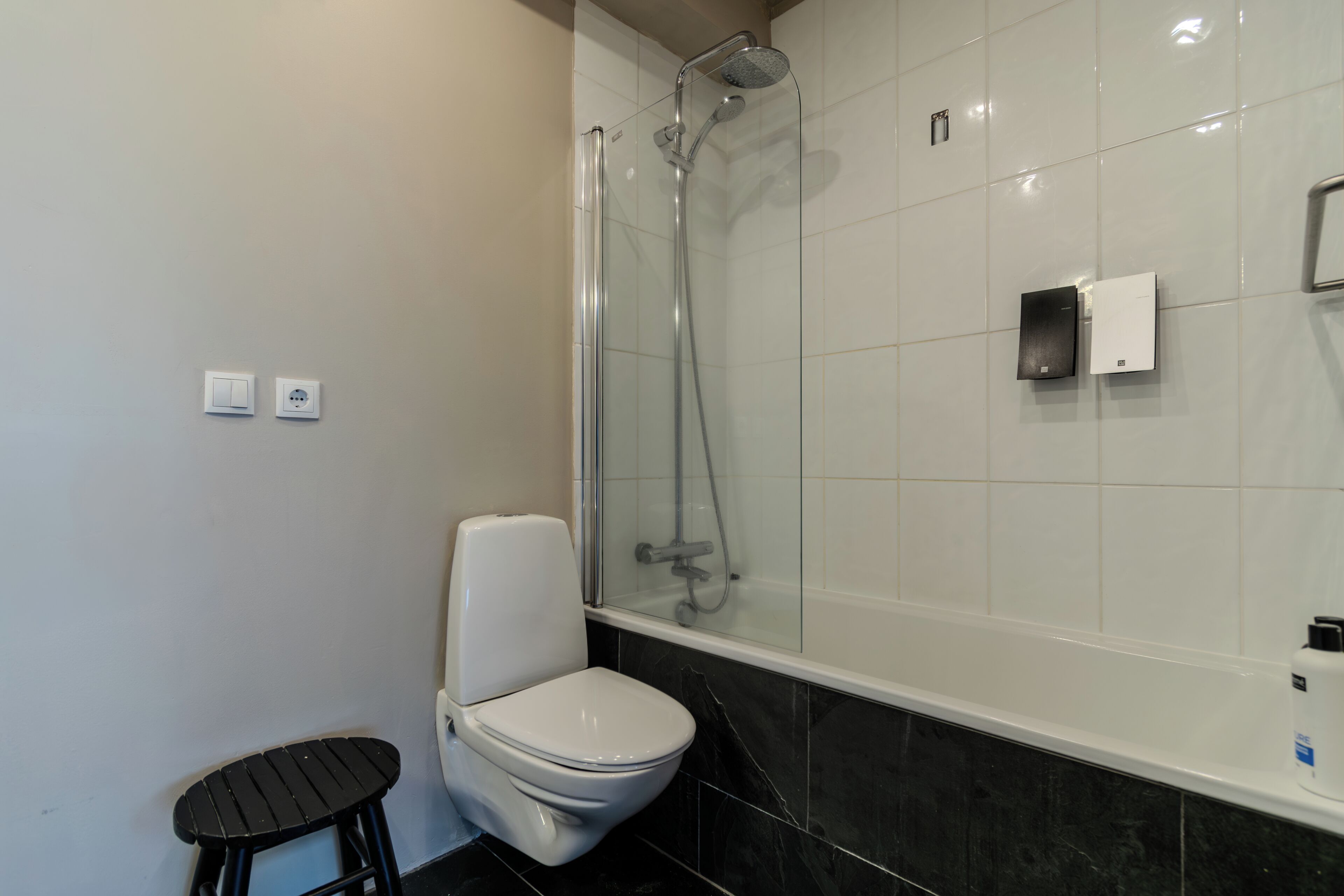 A bathroom with a toilet, a small stool, a shower with a glass door, and a white tiled wall.