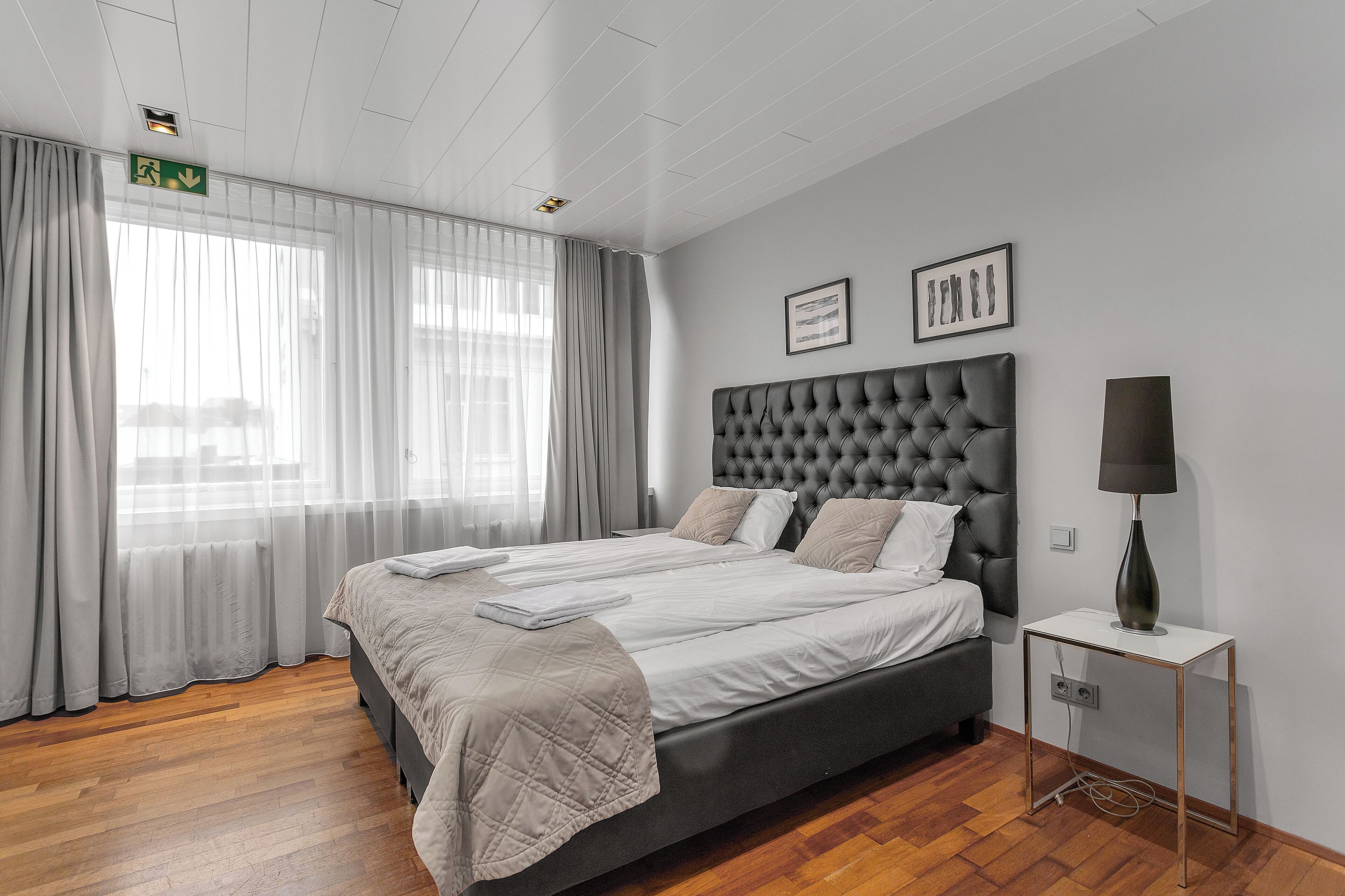 A modern bedroom with a large bed, a bedside table with a lamp, and a view of the outdoors through sheer curtains.