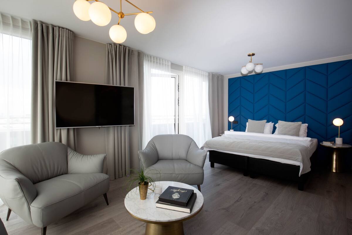 A modern hotel room with a large bed, two armchairs, a TV, and a round coffee table with a book and a plant.