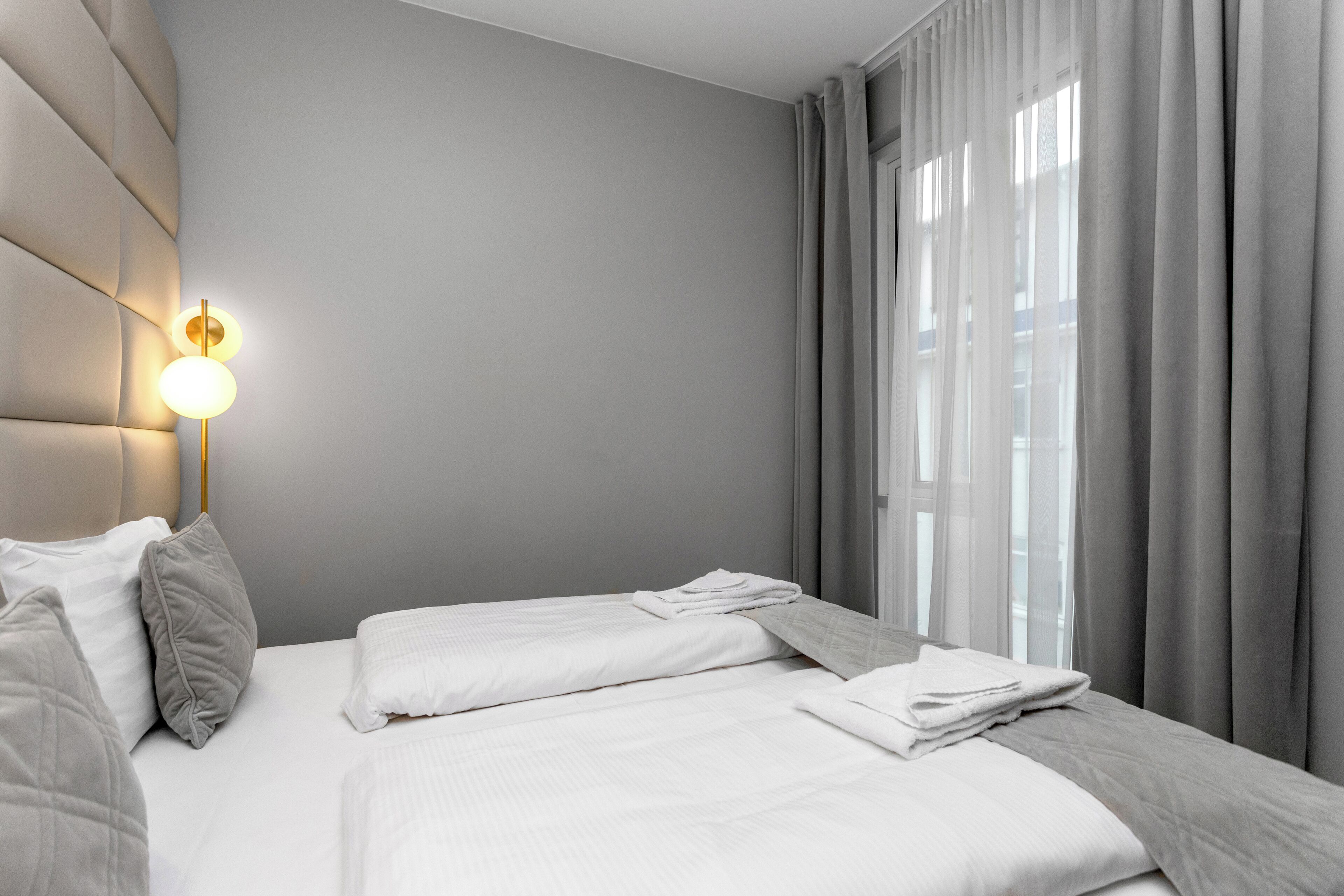 Ein modernes Hotelzimmer mit einem großen Bett, einer Wandleuchte und Blick auf die Stadt durch transparente Vorhänge.