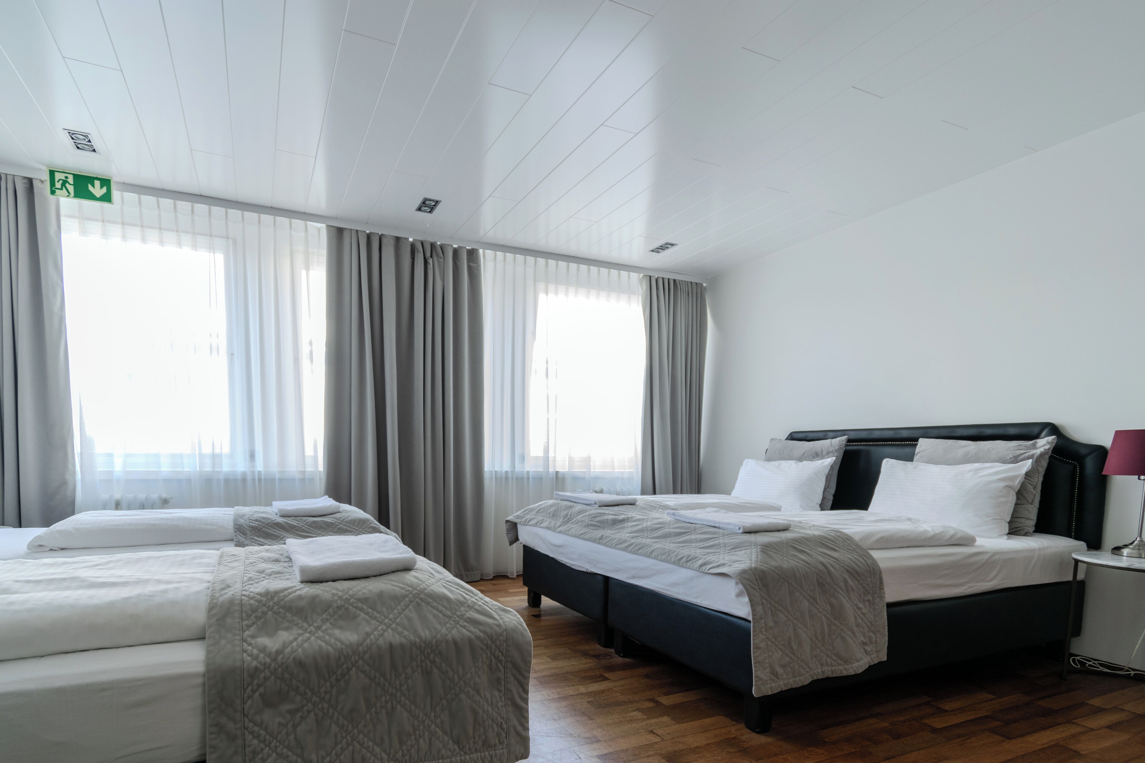 A hotel room with two beds, a wooden floor, a large window with curtains, and a green exit sign.