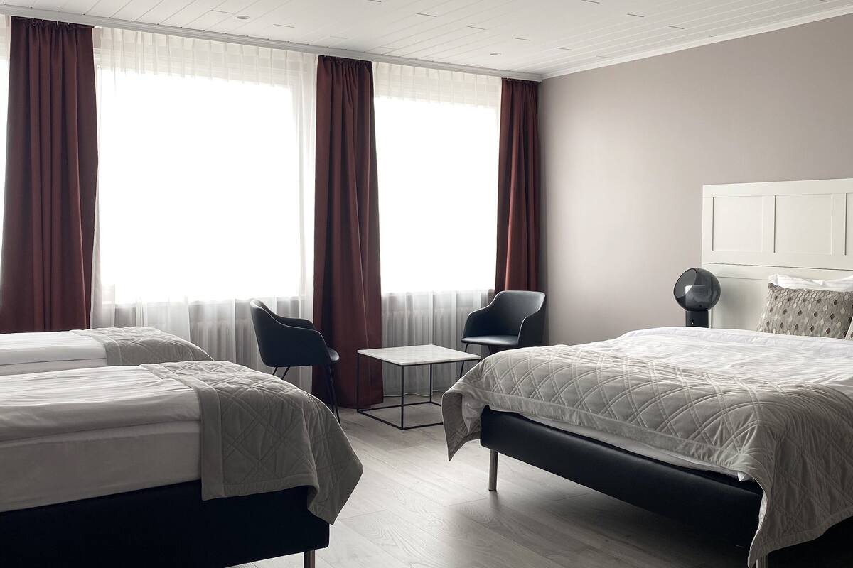 A modern hotel room with two beds, a small table, a chair, and a lamp.
