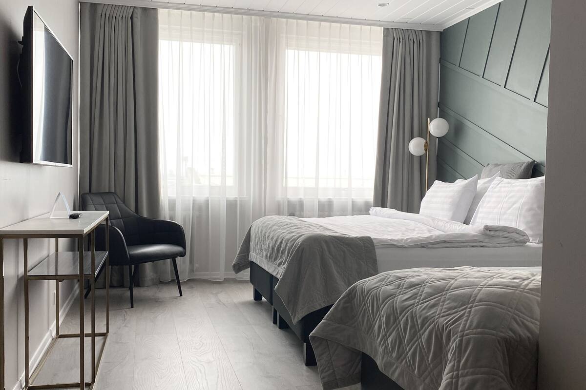 A modern hotel room with a wooden floor, two beds with white bedding, a desk with a chair, a flat-screen TV, and a large window with sheer curtains.