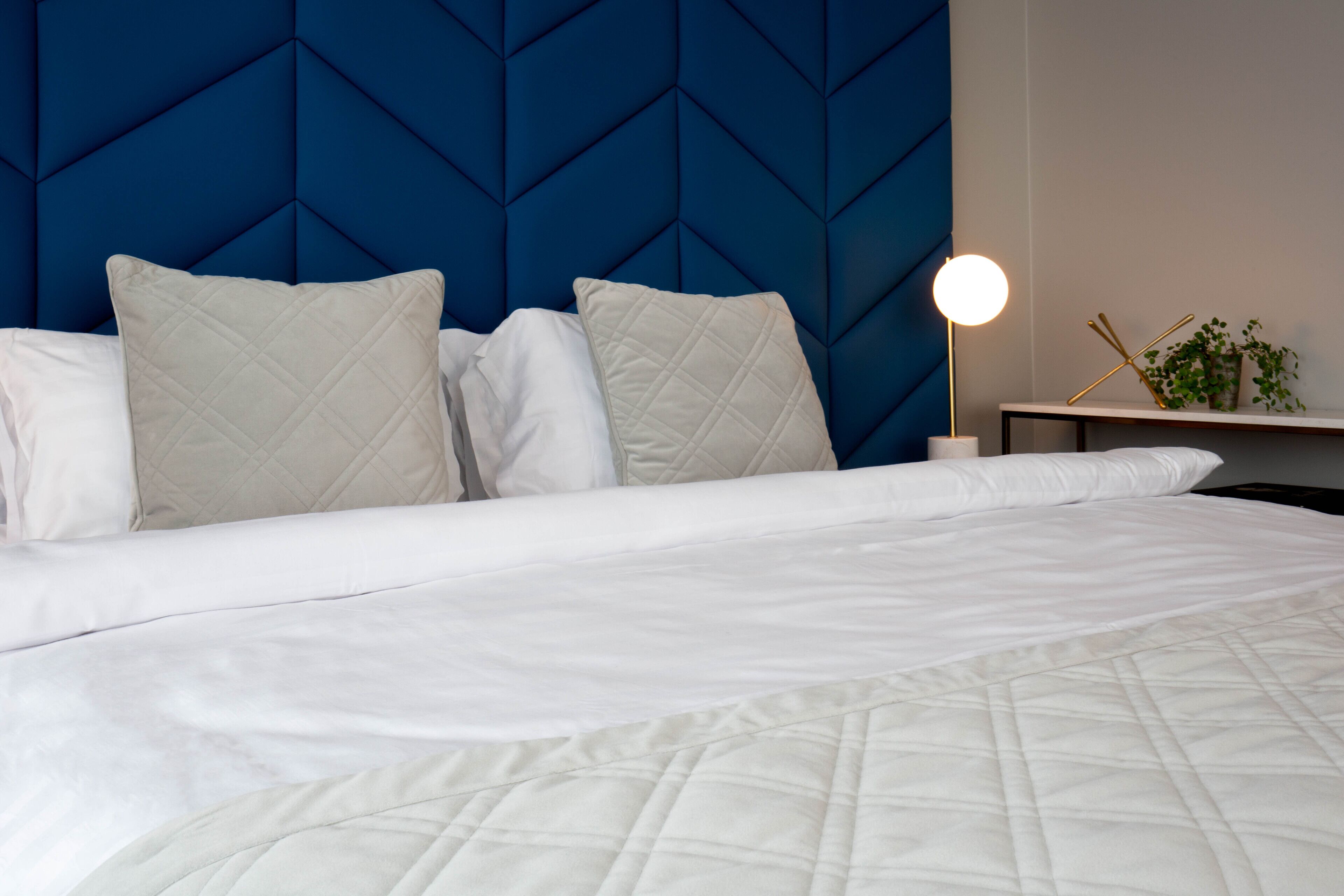A neatly made bed with a quilted white comforter and two pillows, a blue geometric headboard, and a bedside table with a lamp and a plant.