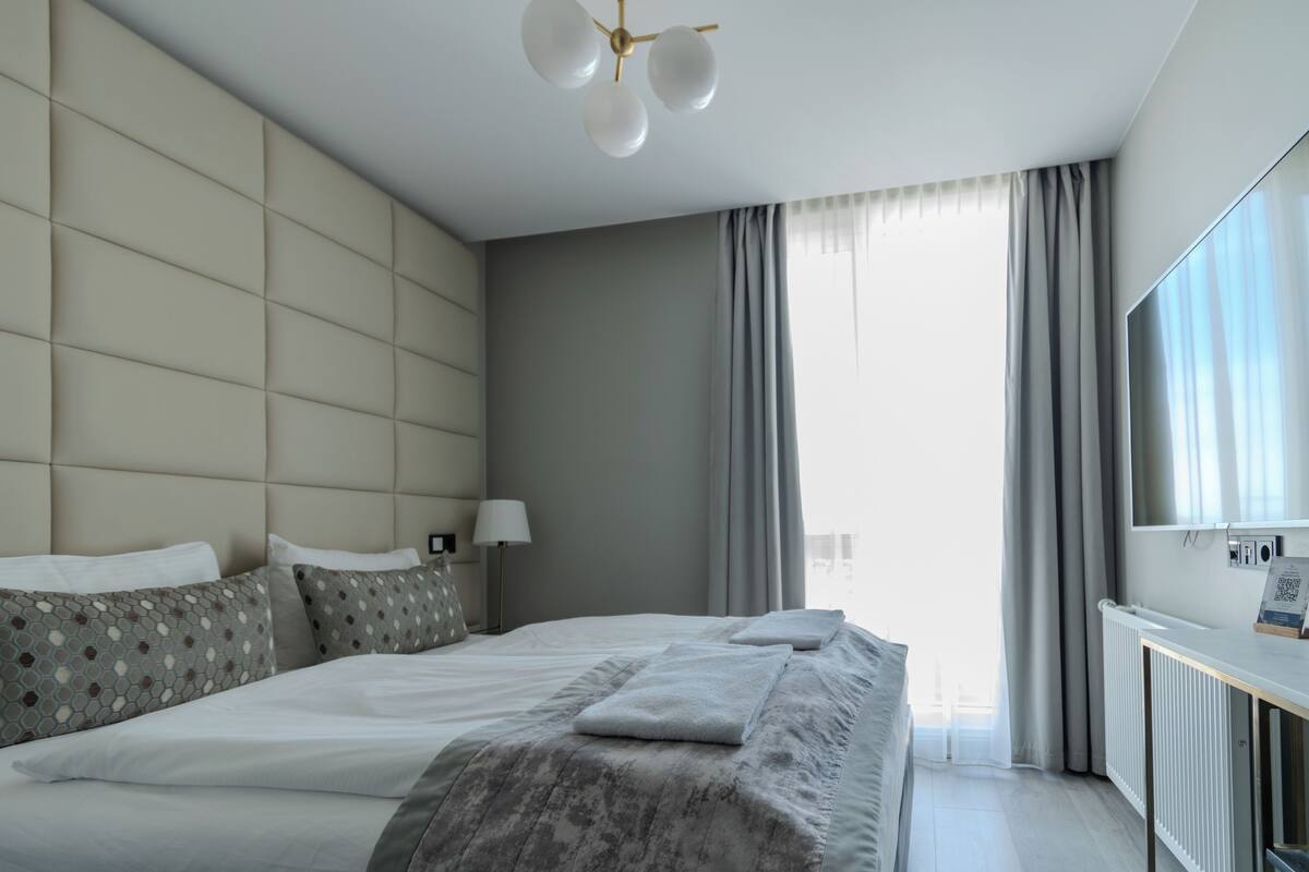 A modern hotel room with a large bed, a bedside table, a mirror, and a view of the city.