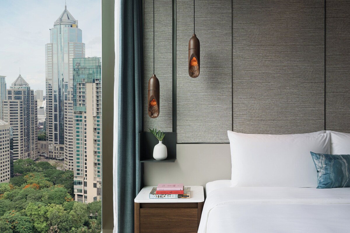 Habitación de hotel moderna con vistas a la ciudad, cama blanca, mesita de noche con libros e iluminación colgante.