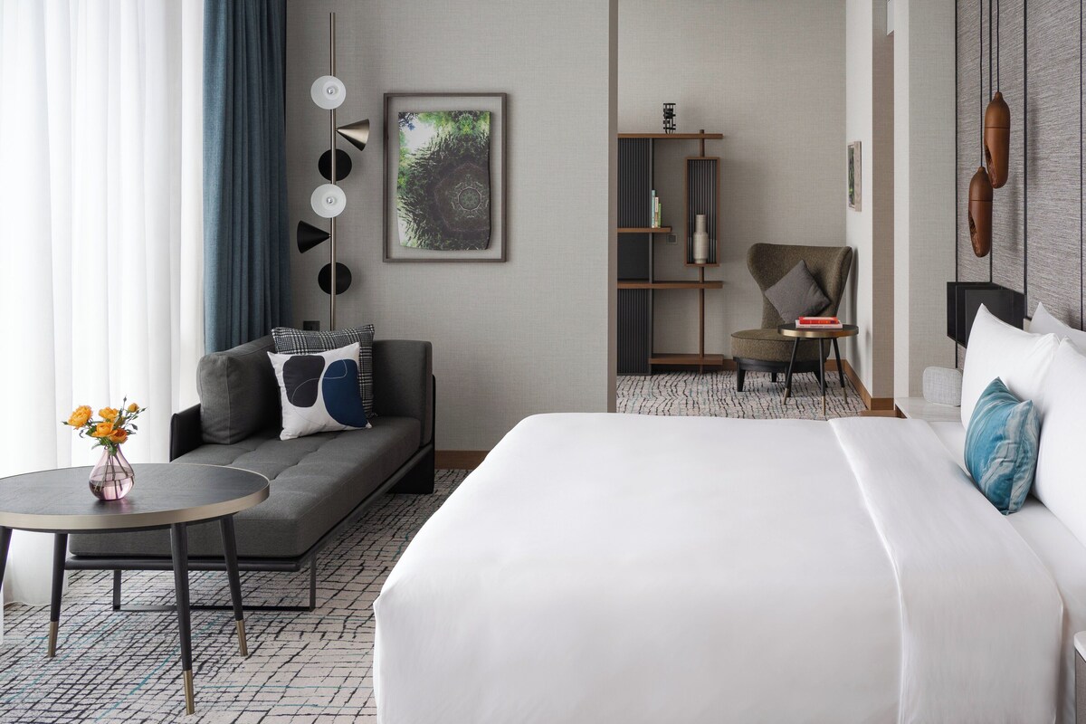 Habitación de hotel con una cama grande, un sofá, una mesita con un florero, una estantería y una silla.