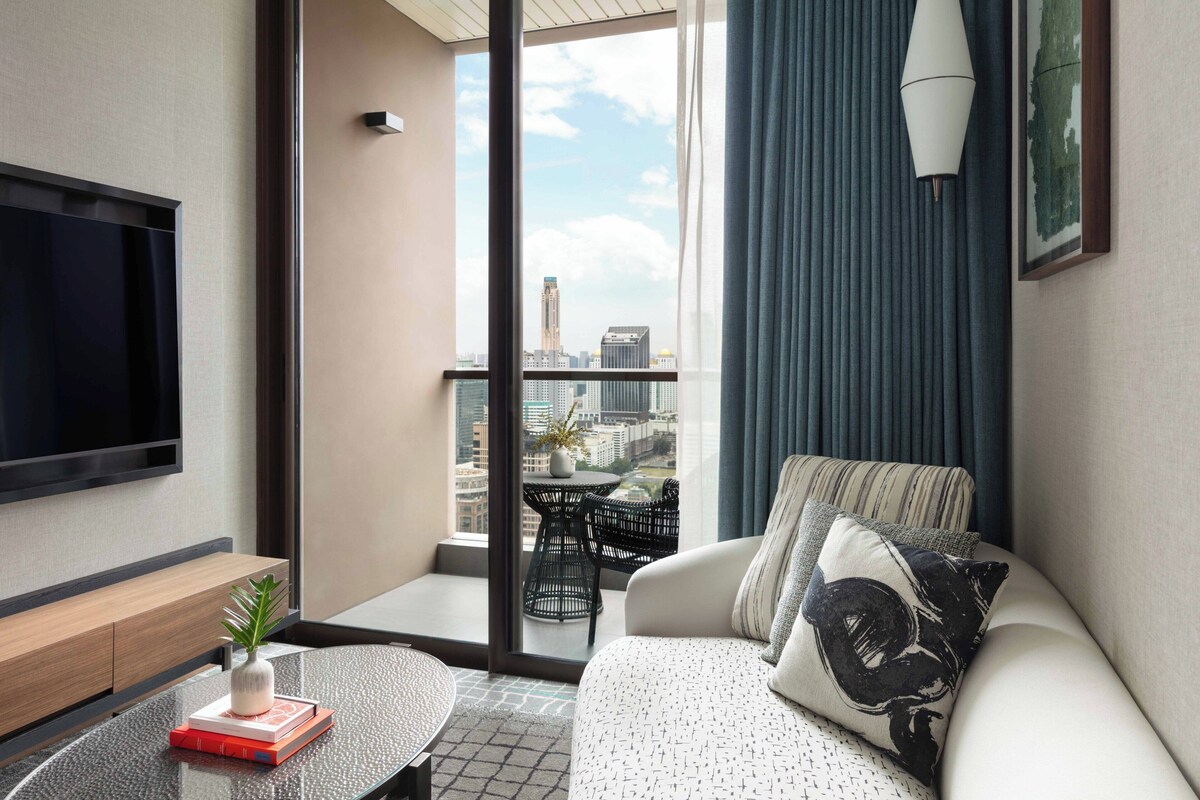 Une chambre d’hôtel moderne dotée d’une grande fenêtre, d’un téléviseur à écran plat, d’un canapé avec des coussins à motifs, d’une petite table agrémentée d’un vase et de livres, et offrant une vue sur le paysage urbain.