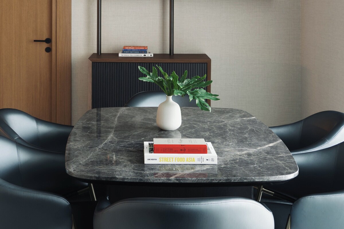 Un comedor moderno con una mesa de mármol, sillas de cuero negro, una puerta de madera, una consola negra, un estante empotrado con libros, un jarrón con hojas verdes y un cuadro abstracto colorido.