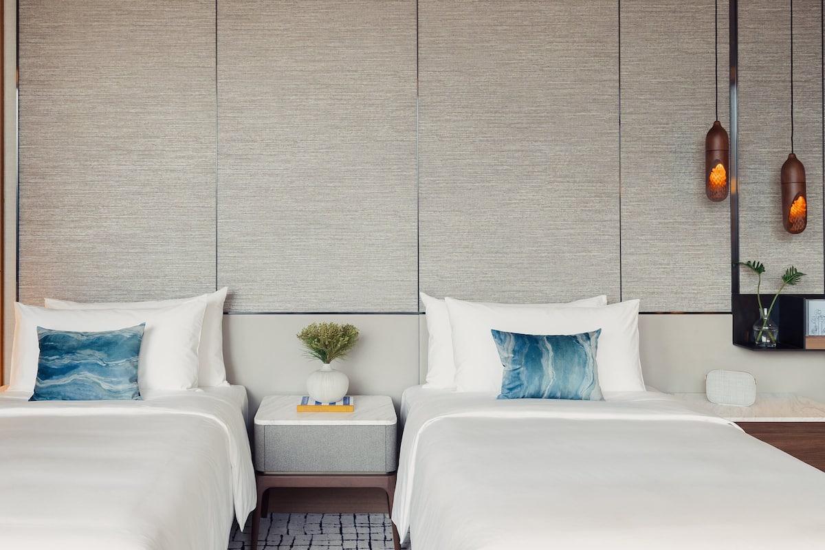 Ein Hotelzimmer mit zwei Betten, einem Nachttisch mit Lampe, einer kleinen Pflanze und einem Spiegel an der Wand.