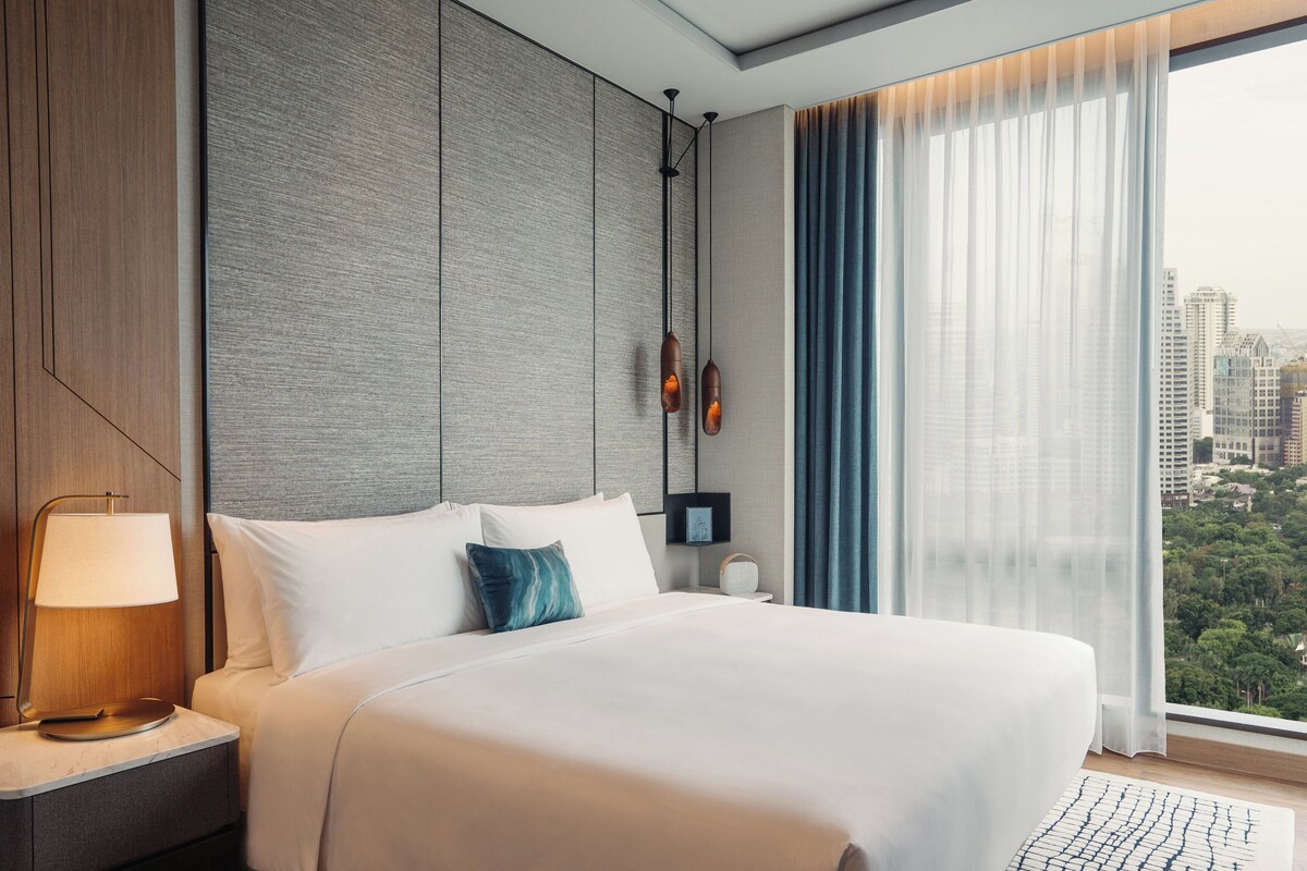 Un hotel moderno con una cama grande, una mesita de noche con lámpara, vistas de la ciudad a través de cortinas traslúcidas, y un paisaje urbano visible por la ventana.