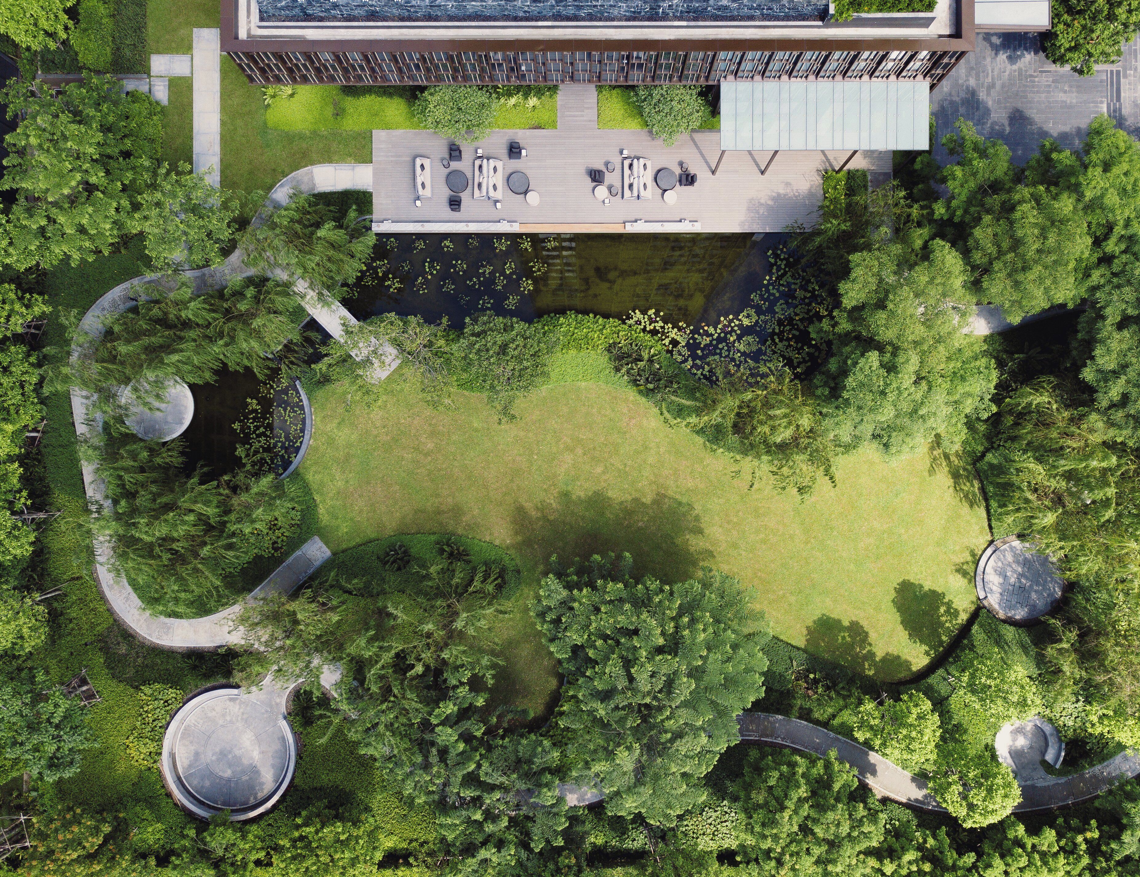 An aerial view of a modern building with a rooftop patio, surrounded by lush greenery and a small pond.