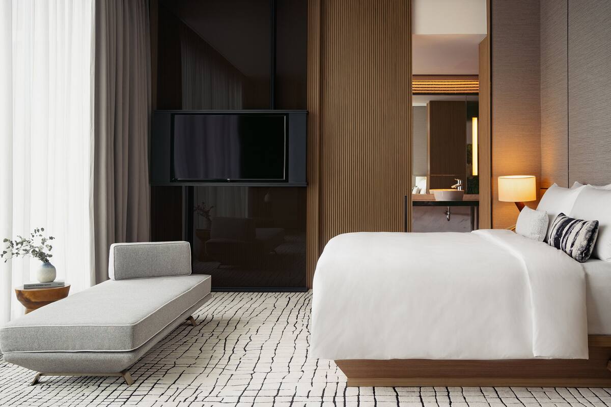 Ein modernes Hotelzimmer mit einem großen Bett, einem Flachbildfernseher, einem Nachttisch mit Lampe und einer kleinen Sitzecke.