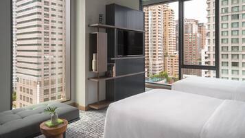A modern hotel room with a large window, a sofa, a bed, and a TV on a shelf.