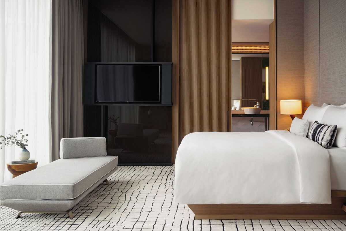 Une chambre d’hôtel moderne dotée d’un grand lit, d’une télévision à écran plat, d’une table de chevet avec une lampe et d’un petit coin salon.