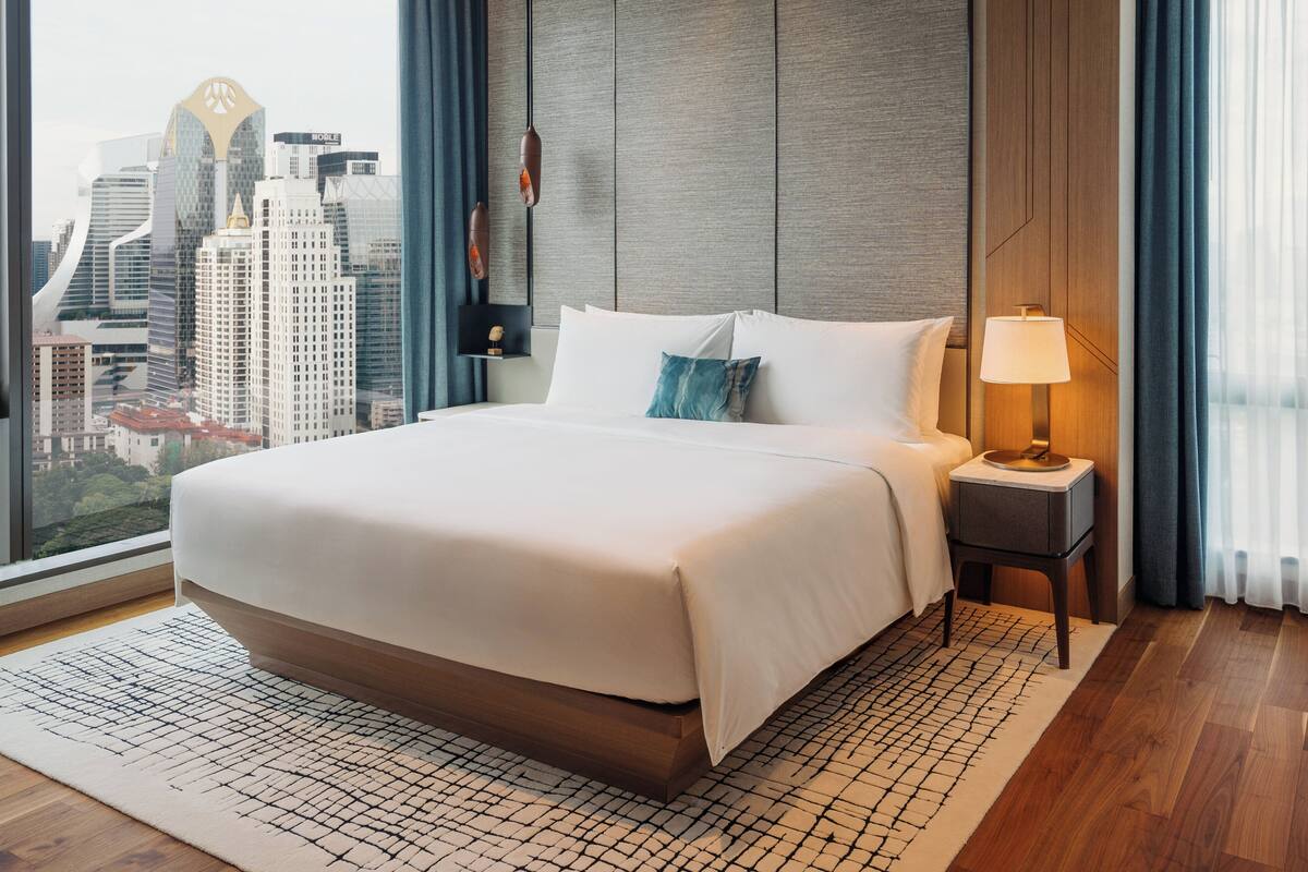 Ein Hotelzimmer mit einem großen Bett, einem Nachttisch mit Lampe, Blick auf die Stadt durch das Fenster und einer Skyline im Hintergrund.