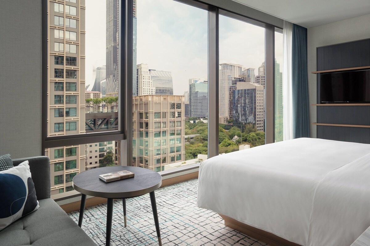 Habitación de hotel con una cama grande, una mesa redonda pequeña, un televisor y vistas a edificios de la ciudad.