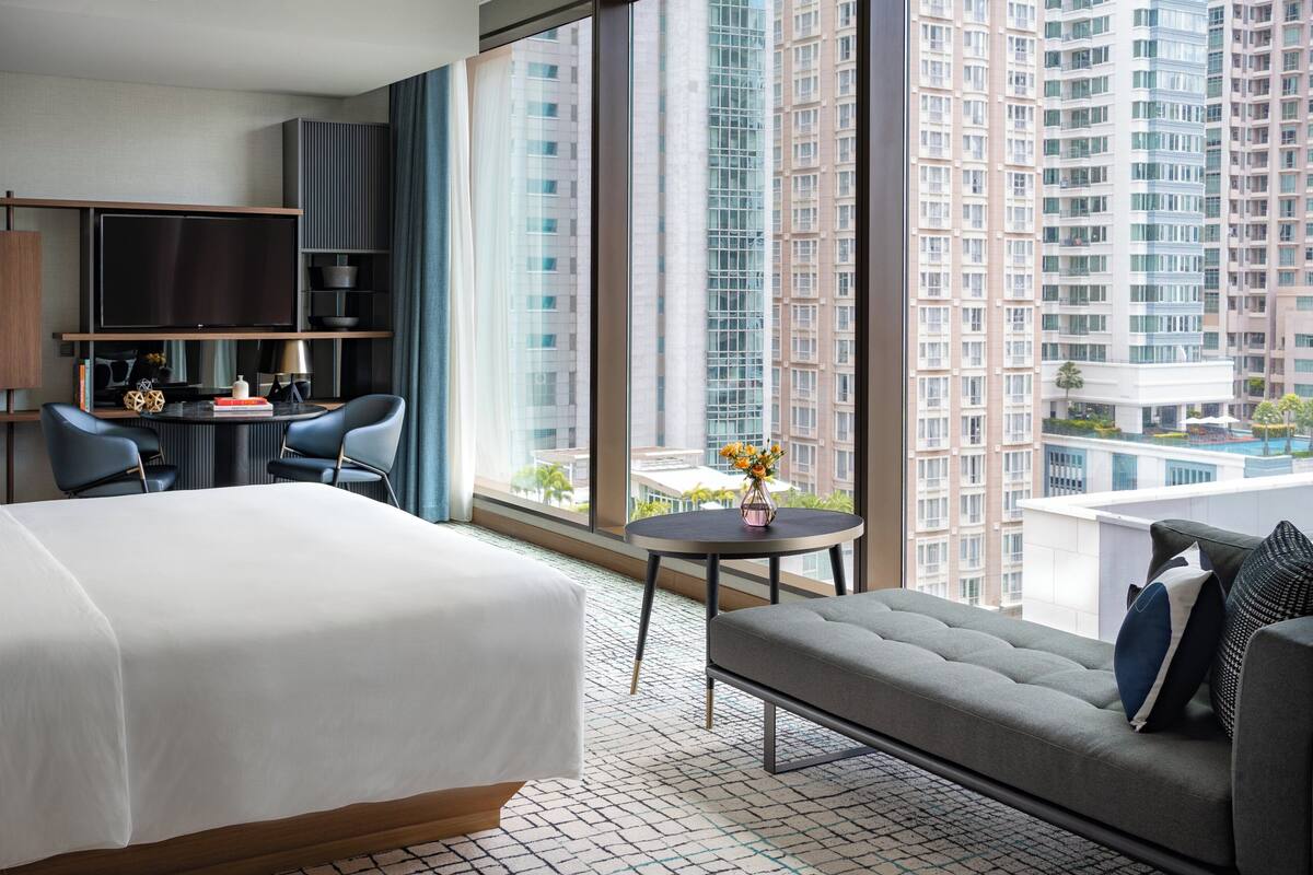 Ein Hotelzimmer mit einem Bett, einer Couch, einem Schreibtisch mit Stuhl, einem Fernseher und einem großen Fenster mit Blick auf die Stadt.