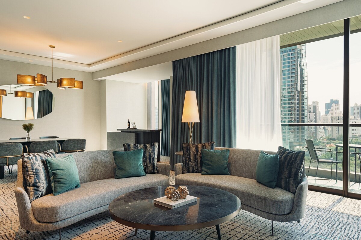 A modern hotel room with a large window offering a city view, a sofa set, a dining table, and a chandelier.