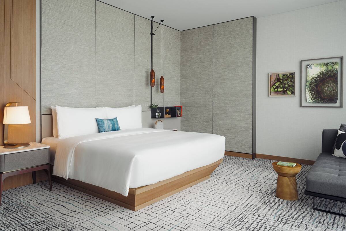 Ein modernes Hotelzimmer mit einem großen Bett, einem Nachttisch mit Lampe, einem kleinen Beistelltisch, einem grauen Sofa und einem gemusterten Teppich.